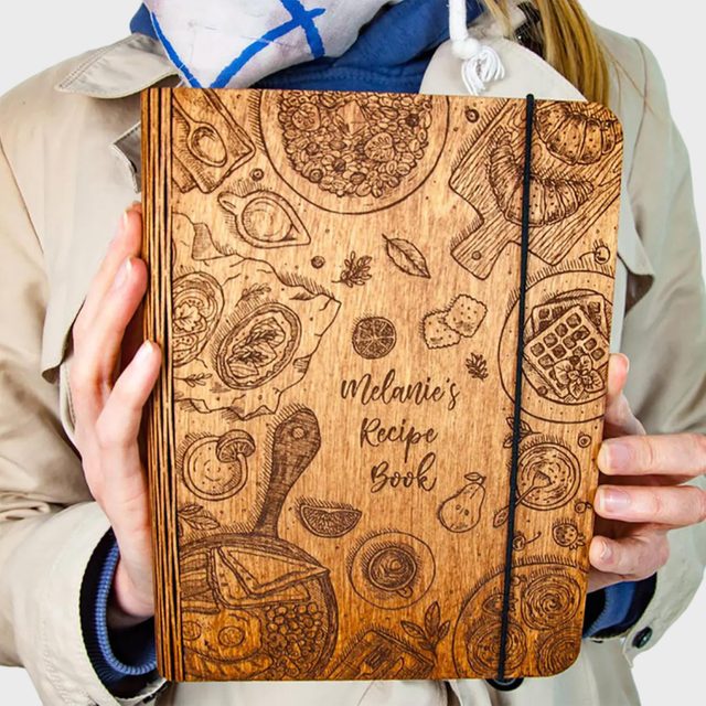 Wooden recipe book with engraved food illustrations held by a person in a coat. Text reads: 