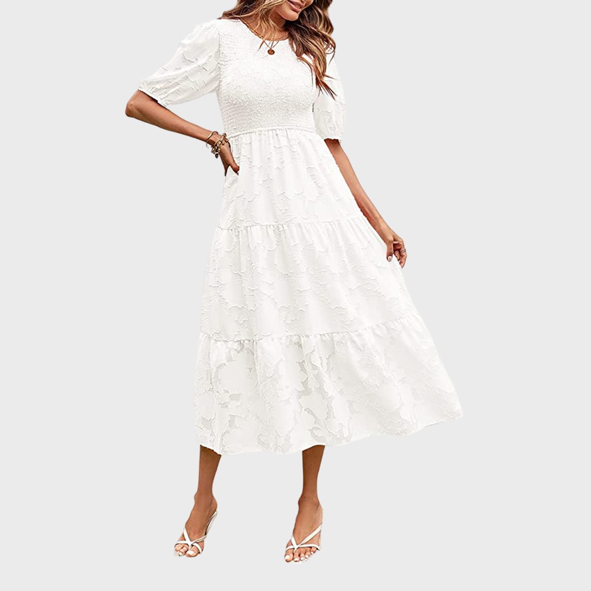 Woman wearing a flowing white dress with puff sleeves, standing in an elegant pose, against a plain light gray background.