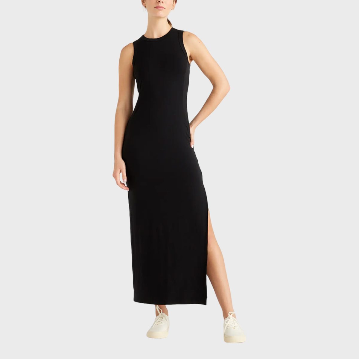 A person stands wearing a sleeveless black dress with a side slit, paired with white sneakers, against a plain gray background.