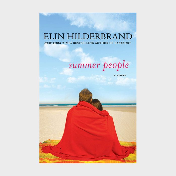 Two people sit wrapped in a red blanket, facing a sandy beach with a clear blue sky. 