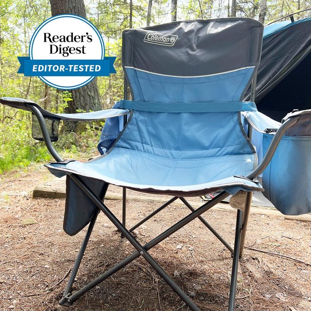 Chair sits unfolded on dirt campsite surrounded by trees; logo reads 