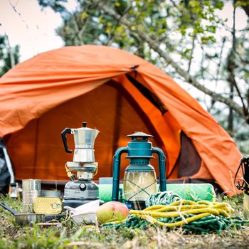Camping gear spread outside an orange tent; includes a coffee maker, lantern, cans, tools, and a rope in a wooded area.