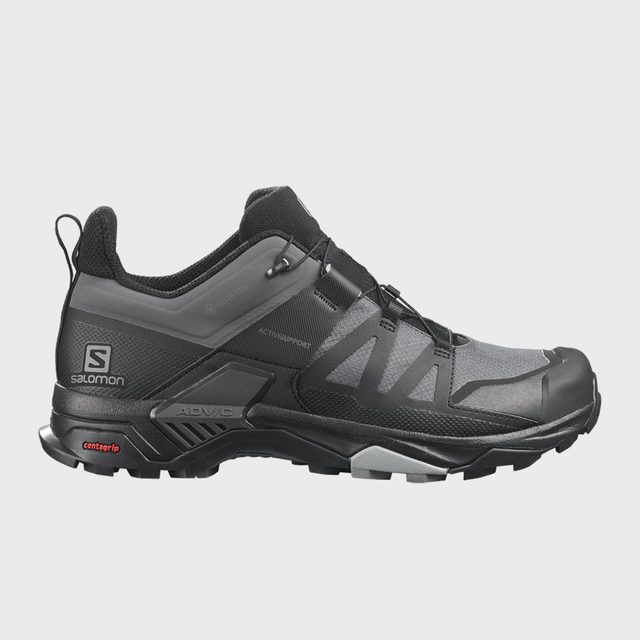 Trail shoe features sturdy design with black and gray tones, visible tread pattern, and Salomon branding. It's designed for durability in rugged terrains.