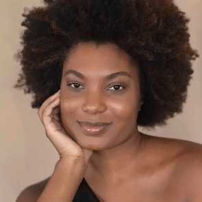 Smiling person rests face on hand; wearing a one-shoulder top. The background is neutral and blurred.