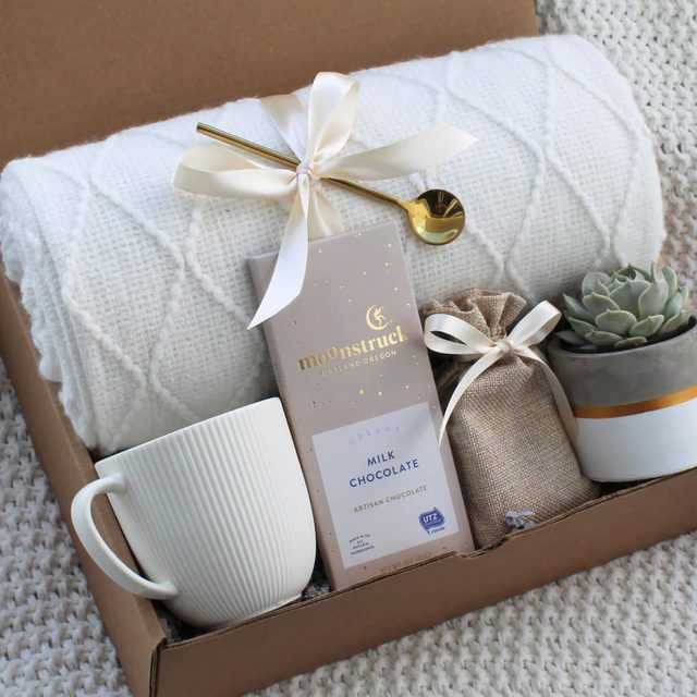 Gift box contains a white blanket, mug, chocolate bar, pouch, golden spoon, and succulent. Items are arranged on textured fabric.