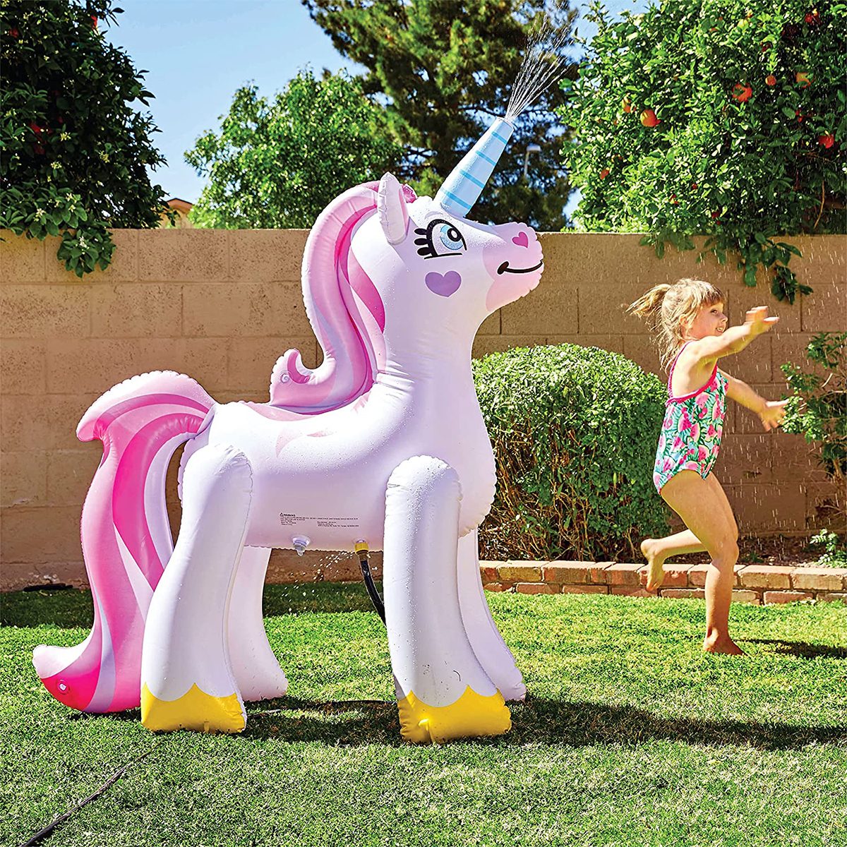 Inflatable unicorn spouts water; child joyfully runs around; backyard with green grass and bushes.
