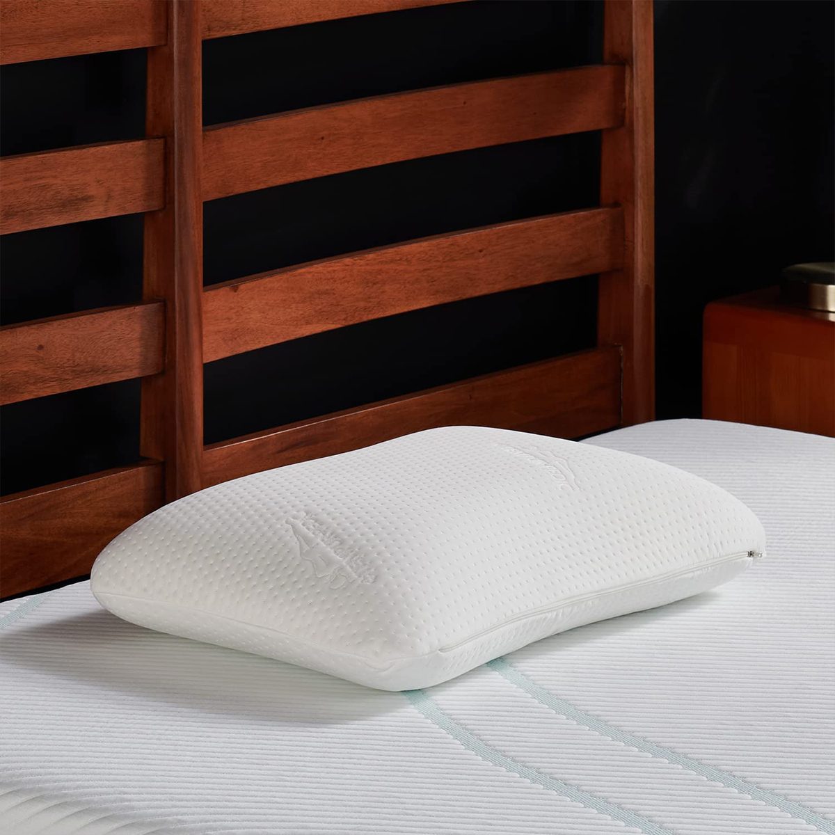 A white pillow rests on a neatly made bed with a wooden slatted headboard in a bedroom setting.