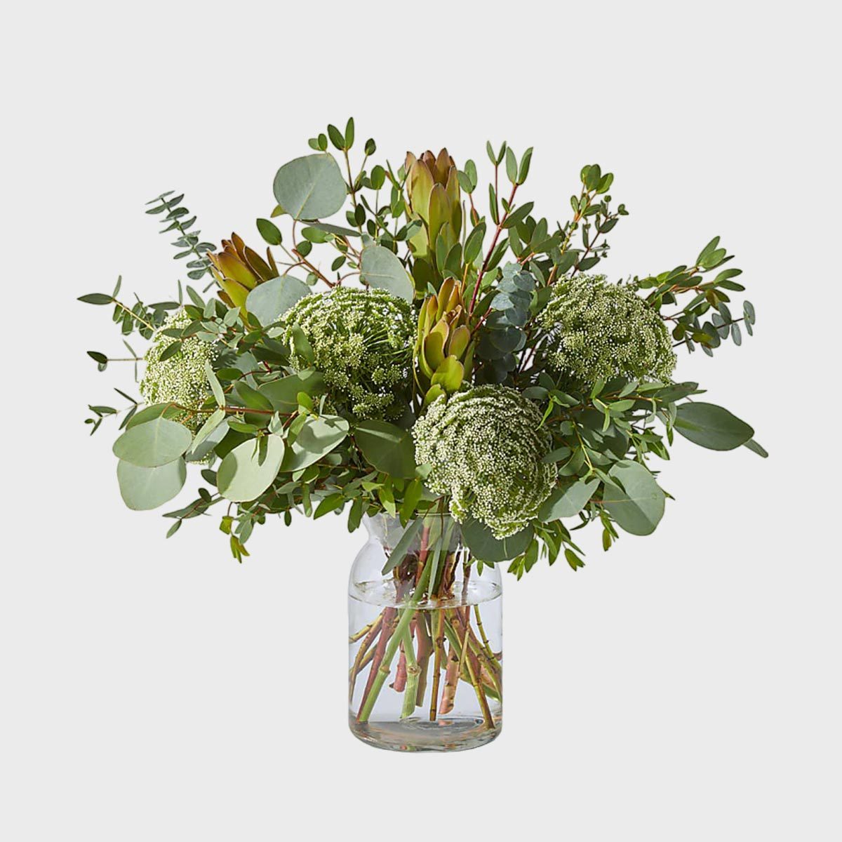 A bouquet of greenery and flowers stands in a clear glass vase filled with water on a plain, light background.