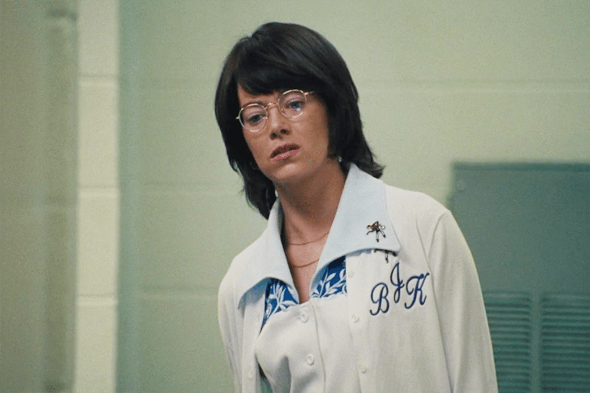 A person wearing glasses and a jacket with "BJK" embroidered stands indoors, against a blurred, green-tinted background.