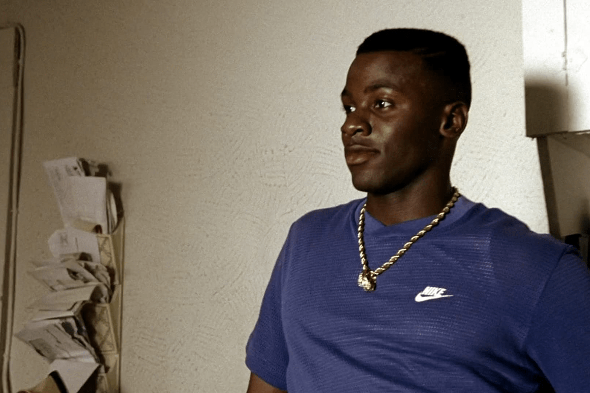 Man wearing blue Nike shirt and gold chain, standing indoors; papers are organized in wall holders beside him.