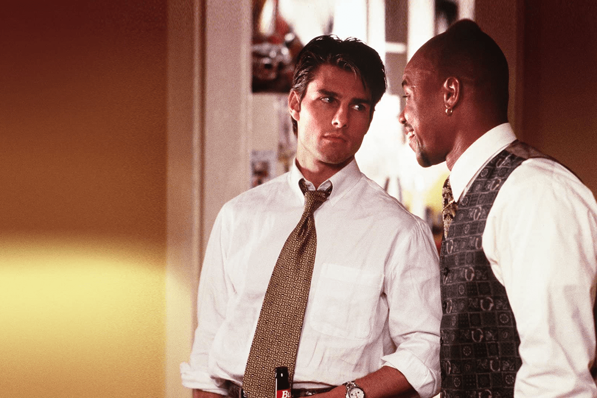 Two men in formal attire converse in a warmly lit room. One holds a Budweiser bottle at waist level.