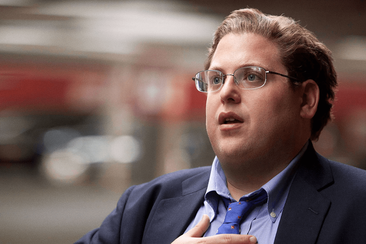 Person talking passionately, hand on chest, in a blurred indoor setting, wearing a suit, blue shirt, and patterned tie.