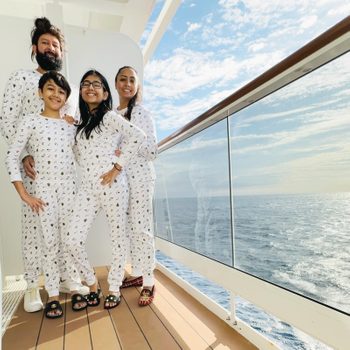 Family smiles in matching pajamas, standing on a cruise ship deck, overlooking the ocean with a clear blue sky.