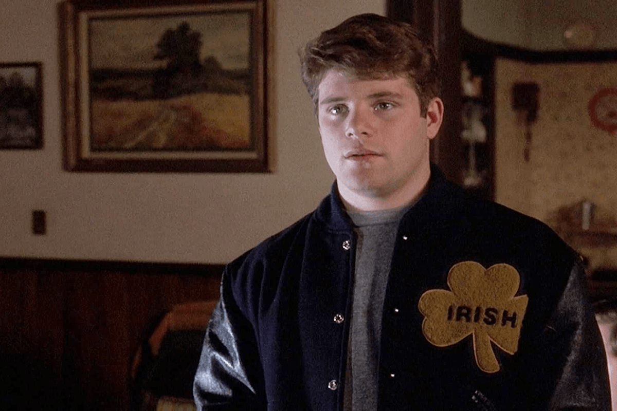 Young man stands wearing a jacket with a shamrock and "IRISH." Indoors, with framed artwork and wood paneling in the background.