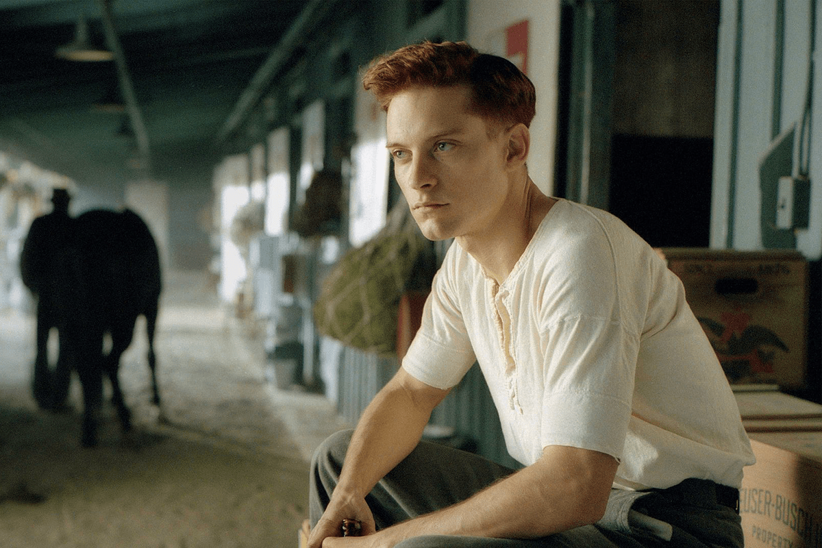 Man sitting intently, wearing a white shirt, in a stable with a horse and person in the background, surrounded by boxes and hay.