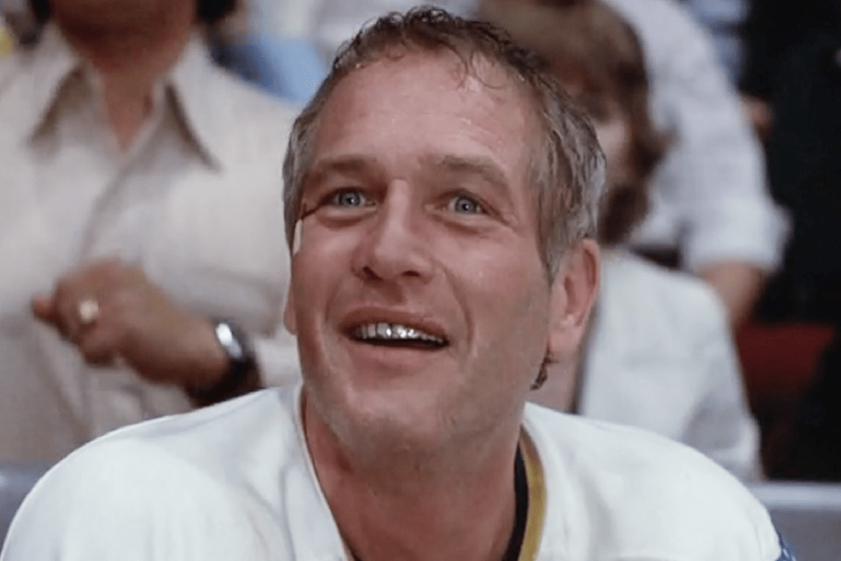 A smiling man with short, light hair looks upward, wearing a white jersey; blurred crowd visible in the background.
