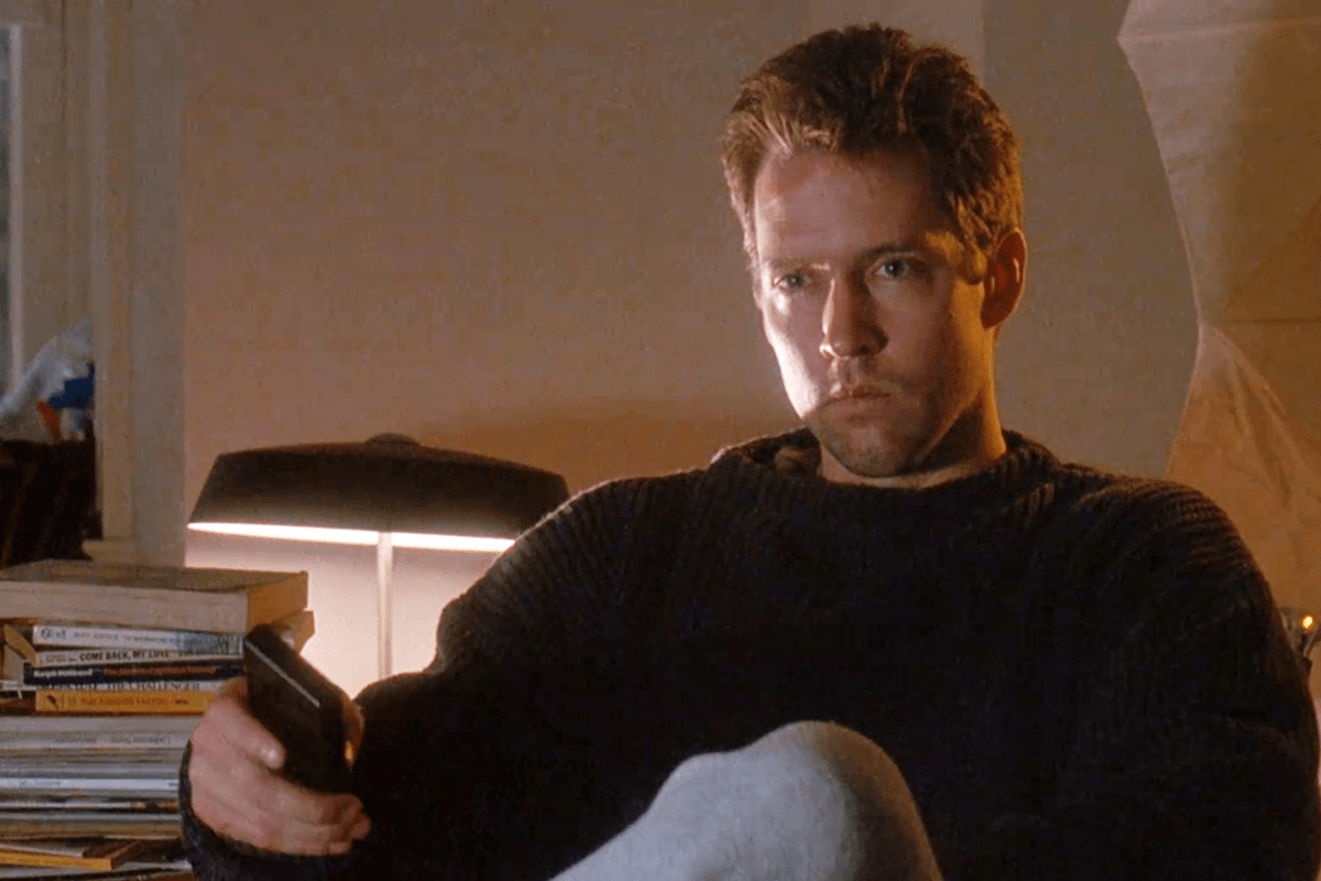 Man holds remote, sitting on couch in dimly lit room with books stacked nearby and a table lamp behind.