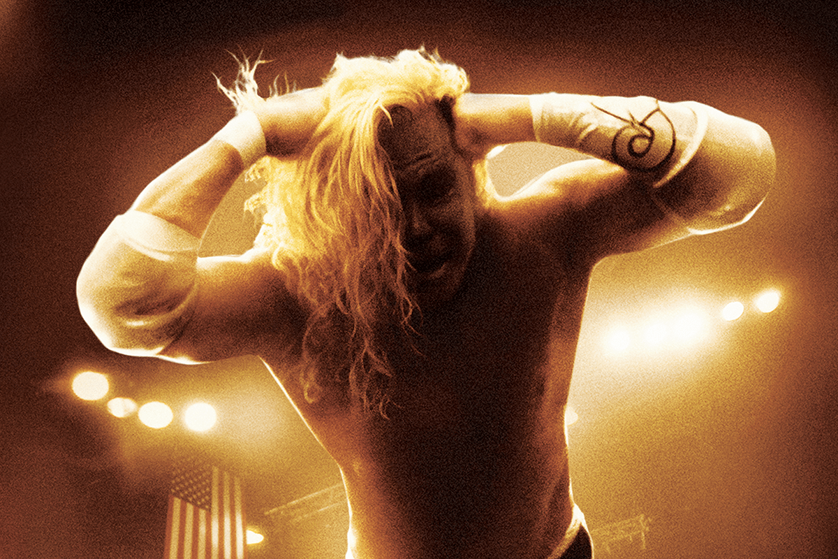 Wrestler clutches head with both hands, hair covering face, in a dimly lit arena with stage lights and an American flag in the background.