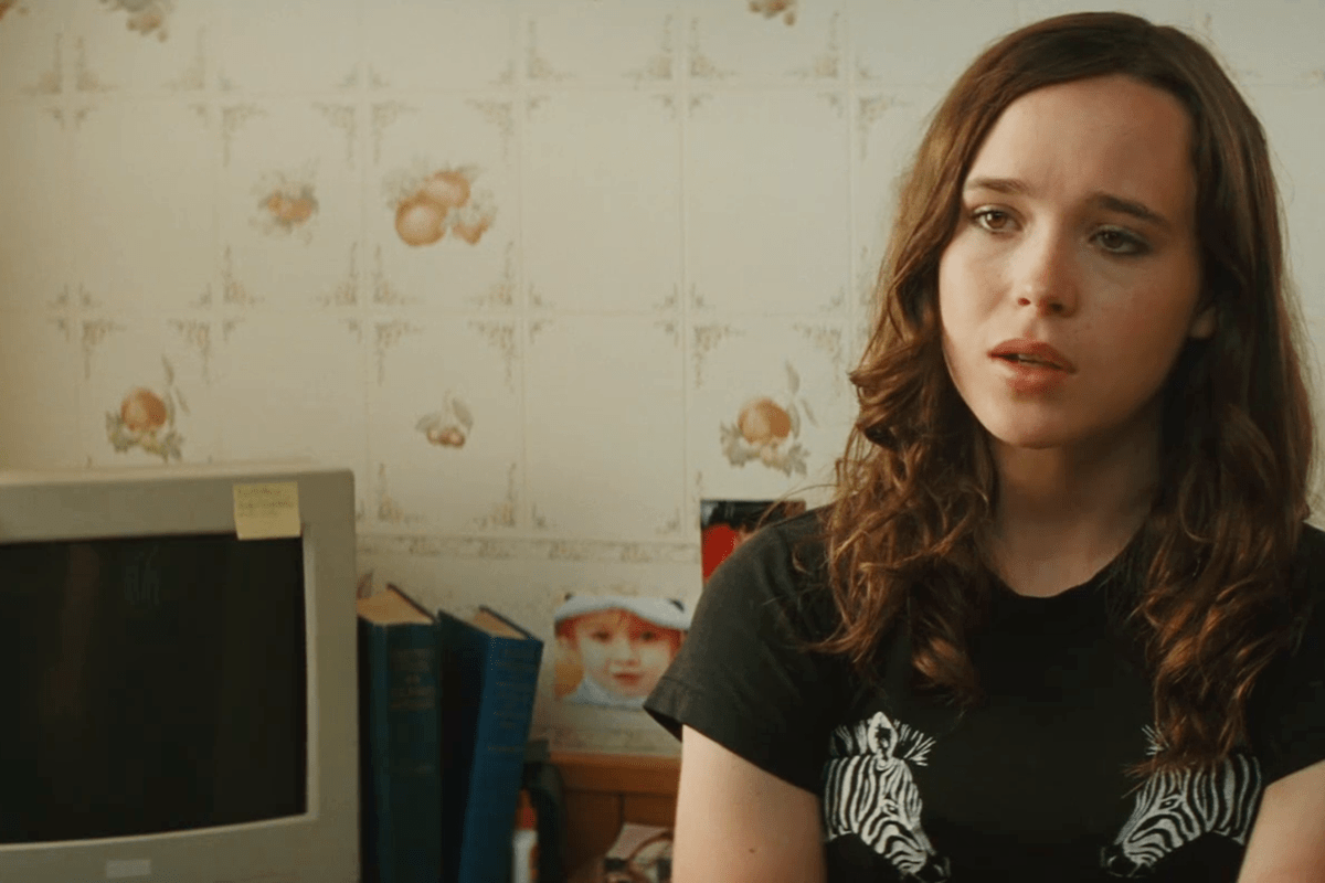 A person sits pensively, wearing a zebra-print shirt in a room with floral wallpaper, next to a computer and books with a sticky note.