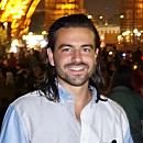 A person smiles, standing outdoors at night, with blurred people and illuminated structures in the background.