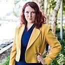 Person wearing a yellow jacket stands confidently outside near trees and greenery, with a blurred background suggesting a park or garden setting.