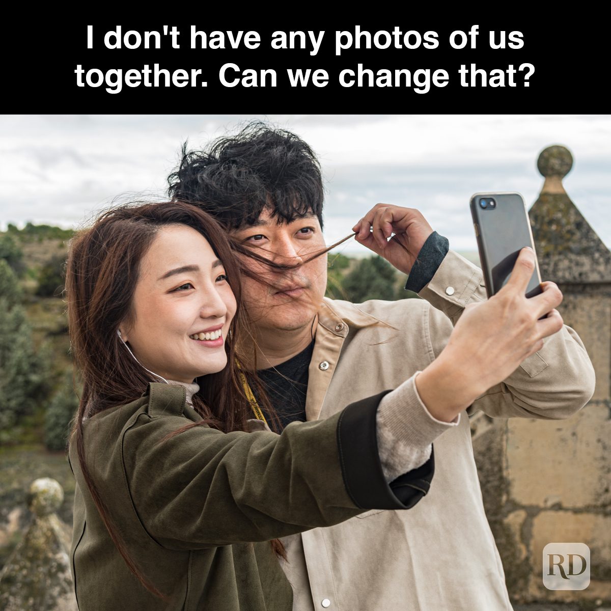People taking a selfie; hair blowing, smiling brightly against a backdrop of stone architecture and trees. Text: "I don't have any photos of us together. Can we change that?"