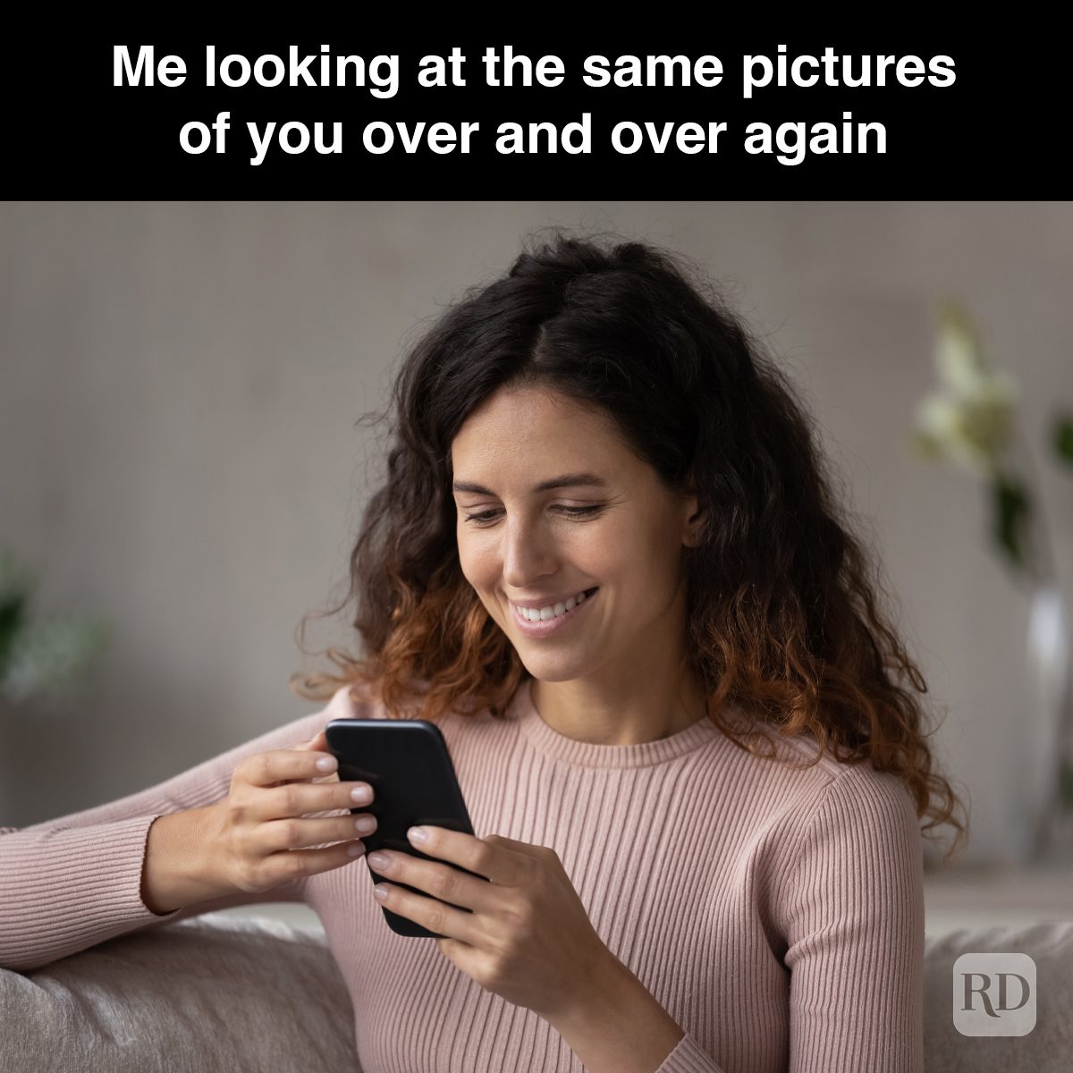 Woman smiling, looking at phone, sitting on a couch with a vase in the background. Text: "Me looking at the same pictures of you over and over again."