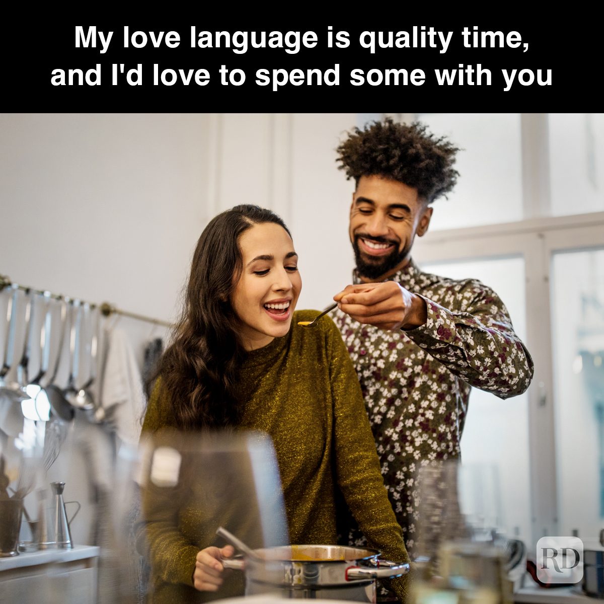 Man offers woman a taste from a spoon, both smiling in a kitchen. Text: "My love language is quality time, and I'd love to spend some with you."
