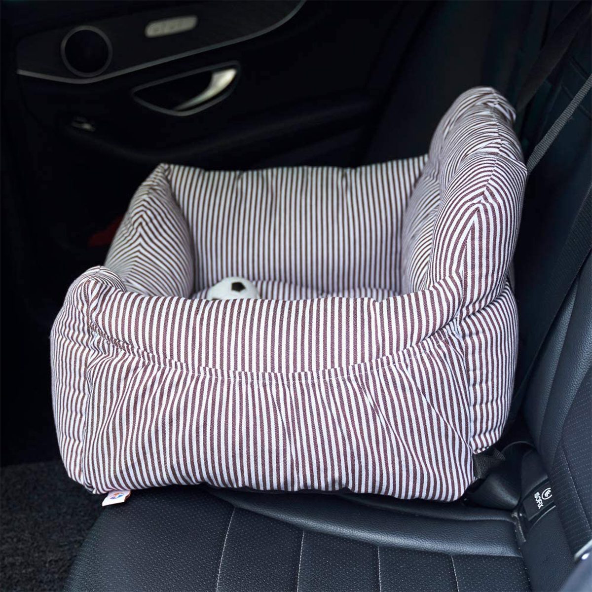 A striped, cushioned pet seat rests on a car's back seat, secured by the seatbelt, with a small toy inside.