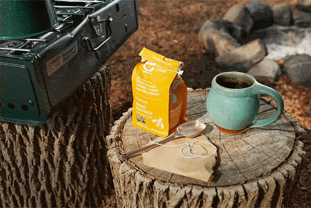 Coffee package and teal mug sit on a tree stump near a campfire pit; adjacent green camping stove visible. Text: "Coffee, sweet espresso."