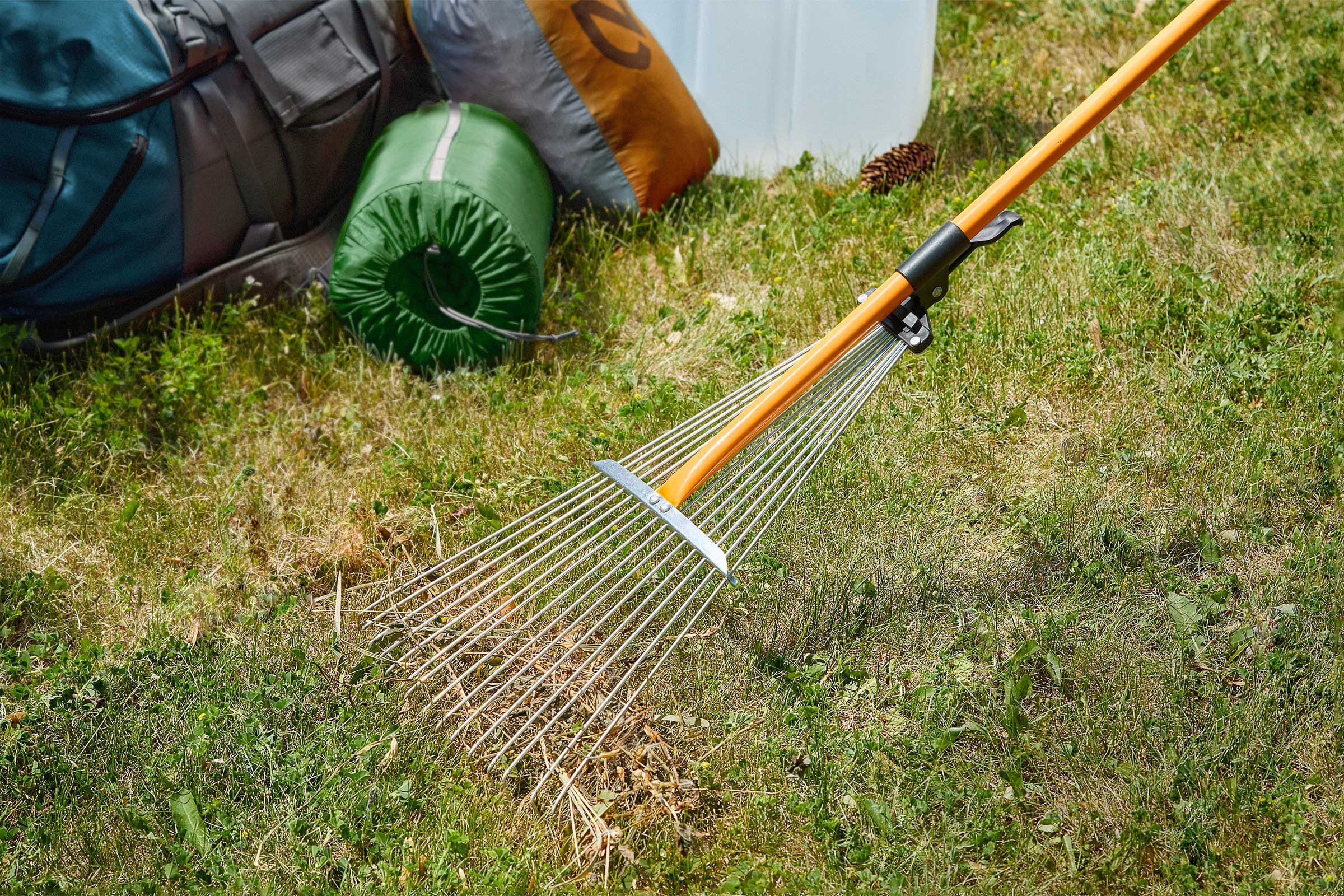 A metal rake rests on grass beside camping gear, including backpacks and a rolled sleeping bag.