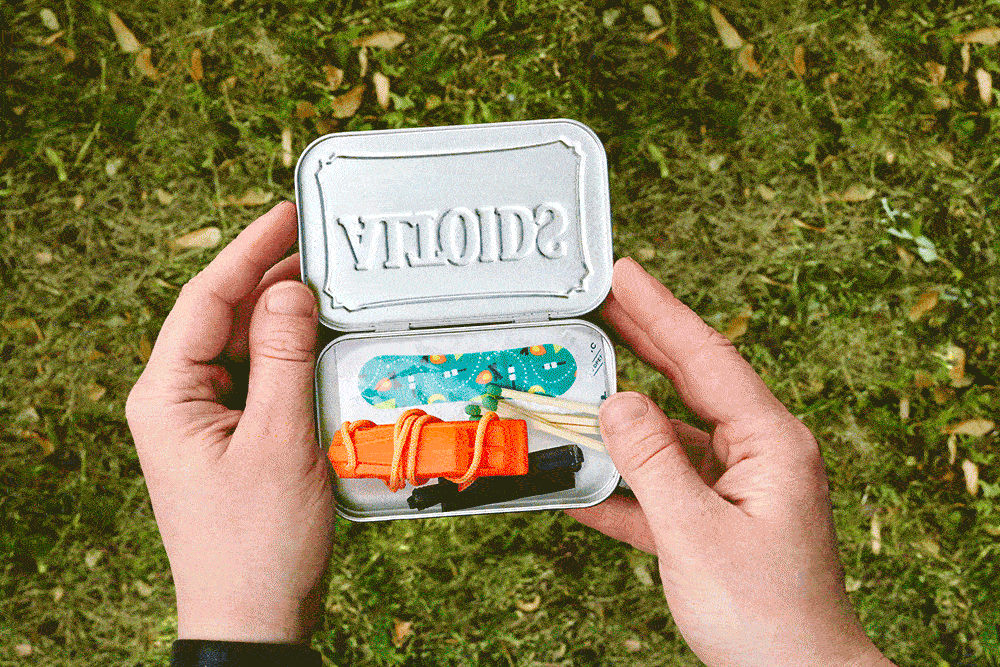 Hands hold an open mint tin containing various survival items, viewed against grassy ground.