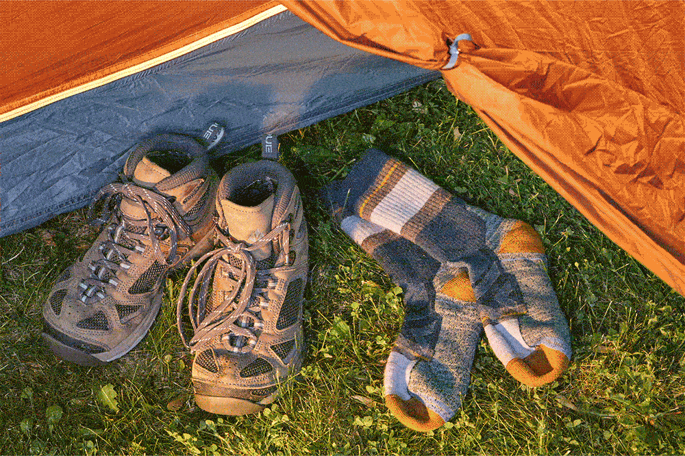 Hiking boots and patterned socks resting on grass outside an orange tent.