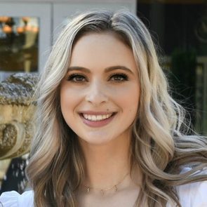 Smiling woman with long, wavy hair stands outdoors, fountain and building in the background.