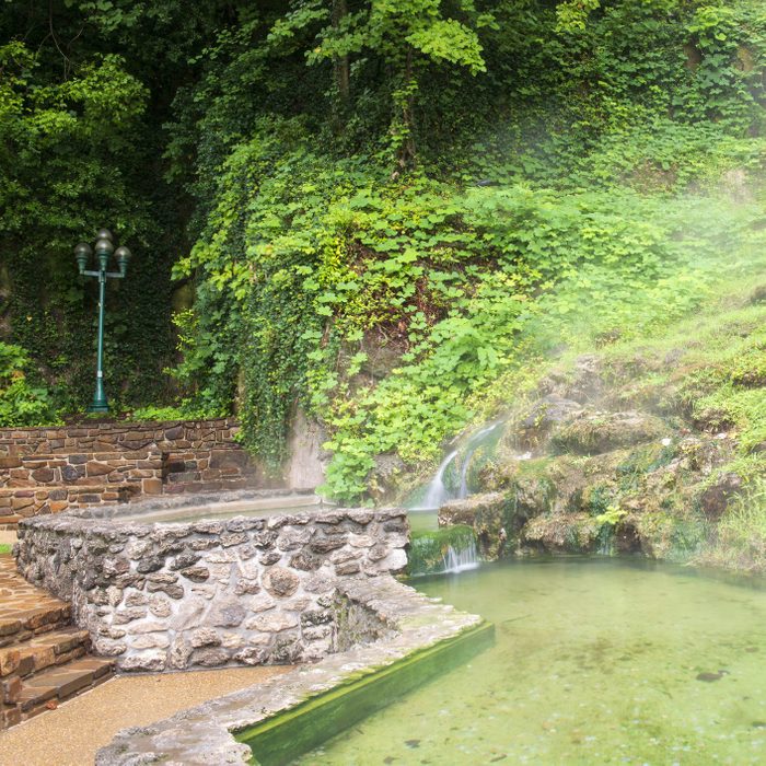 Stone platform with water droplets forming mist, surrounded by lush greenery and a small waterfall, adjacent to a lamppost and stone pathway.