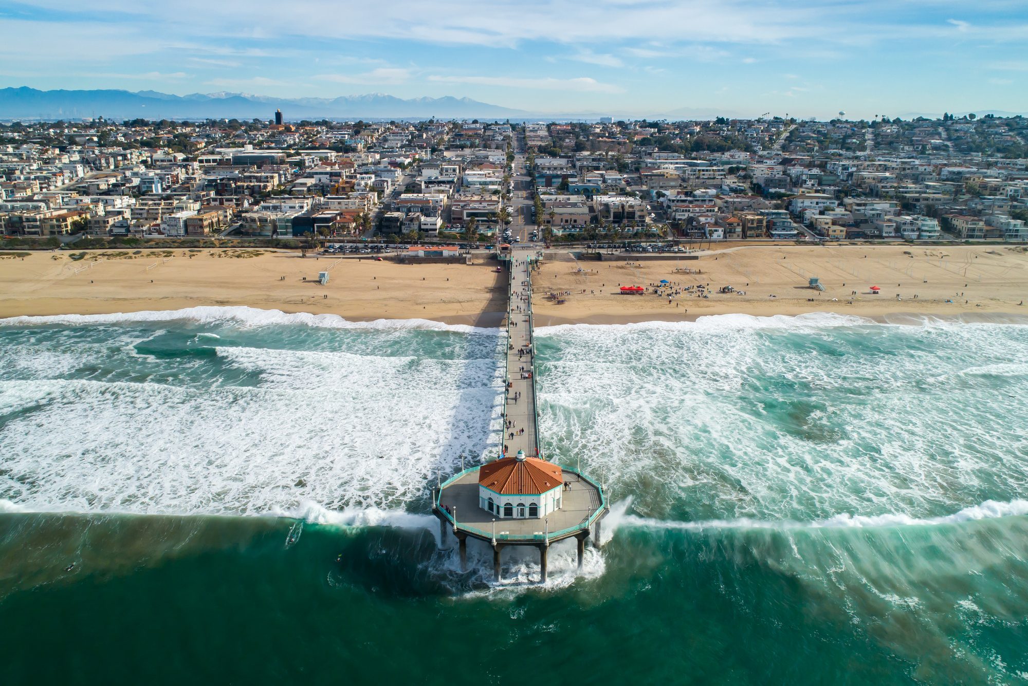 Pier extends over ocean waves, leading from bustling beachfront cityscape under clear skies.