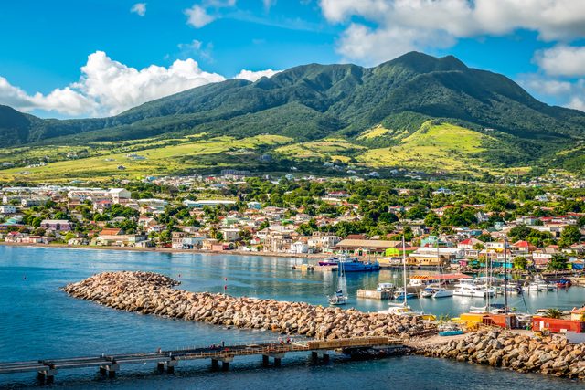Buildings rest by a calm bay, surrounded by vibrant hills and mountains, boats moored, under a bright sky with scattered clouds.
