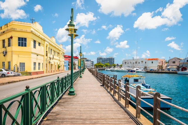 Pier extends toward buildings, lined with green lampposts and railings; boats are docked on the water under a bright blue sky with scattered clouds.