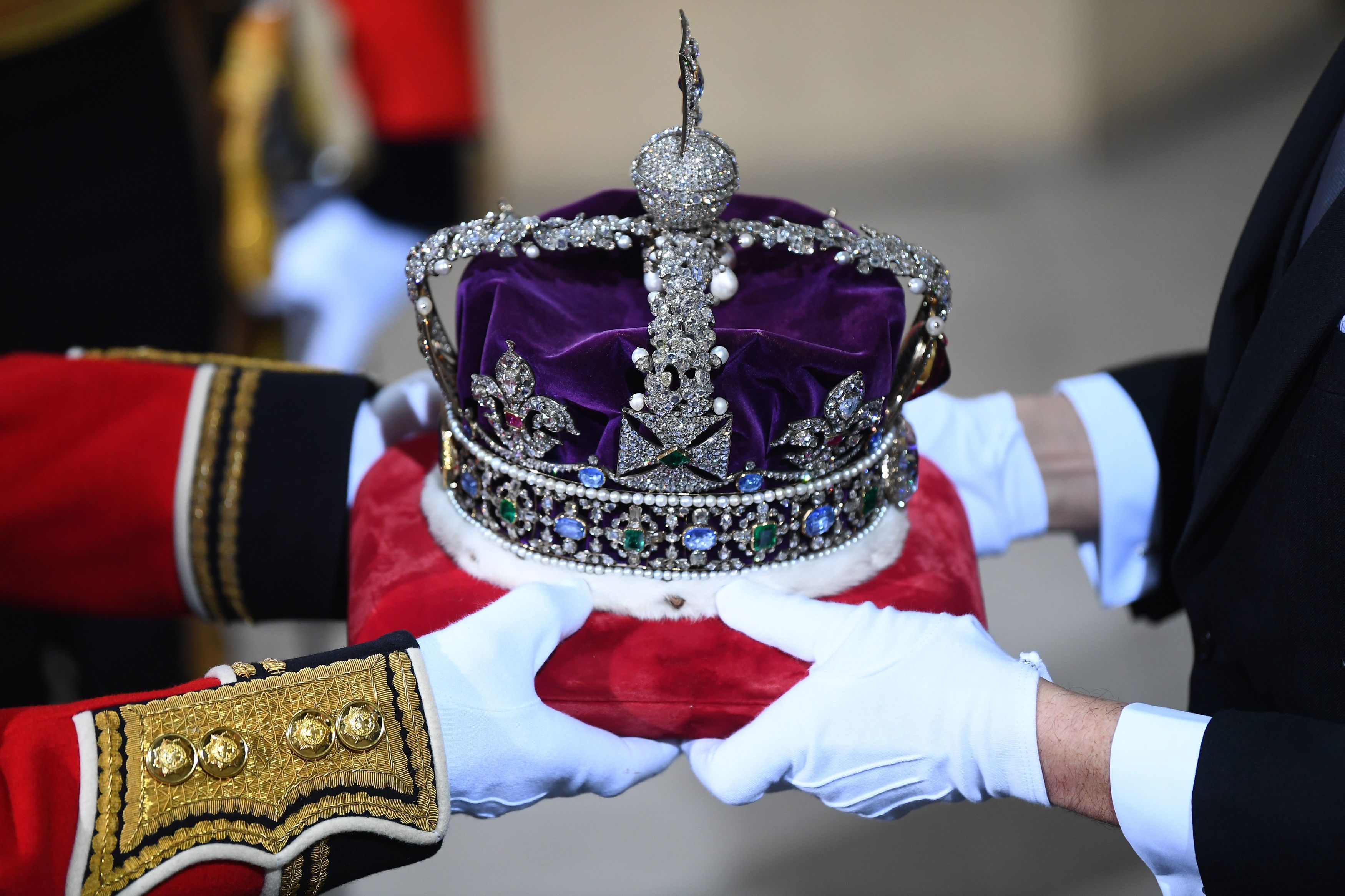 A jeweled crown is being carefully held by two people wearing white gloves, surrounded by formal attire.