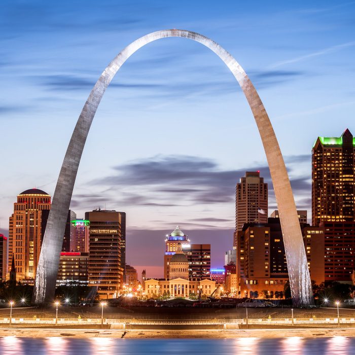 Arch towering over illuminated cityscape at dusk, with a clear sky and vibrant lights reflecting on a river in the foreground.