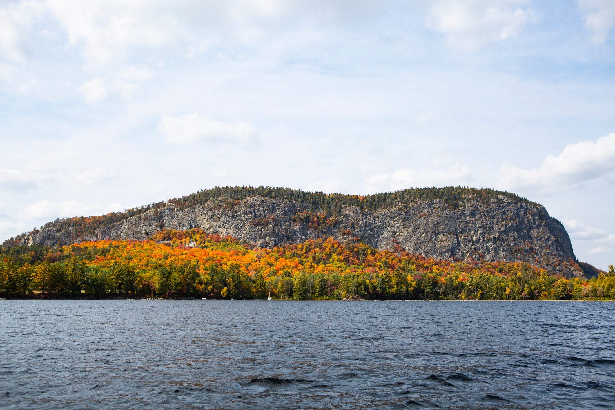 Cliff rises majestically, surrounded by vibrant autumn trees, overlooking a serene lake under a partly cloudy sky.