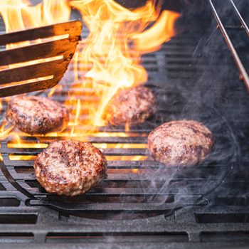 Burger patties grill amid flames, with a spatula flipping one. The smoky barbecue setting heightens the cooking intensity.