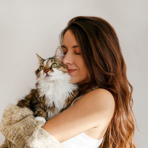 Cat is being cuddled by a woman with long hair in a cozy, neutral-toned setting. The cat looks content and alert.