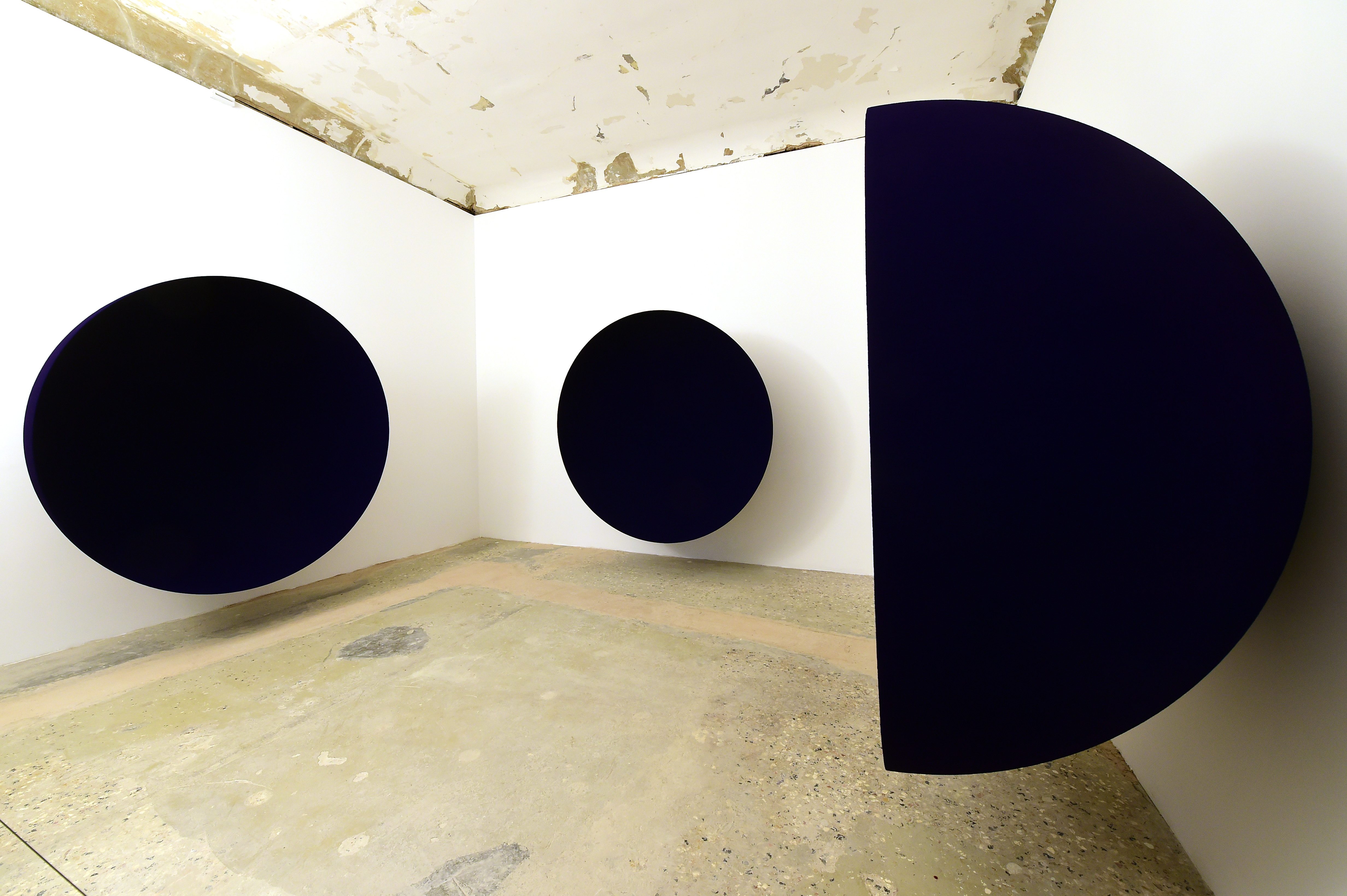 Dark circular shapes appear to float on white walls in a minimalist, unfinished room with a textured concrete floor.
