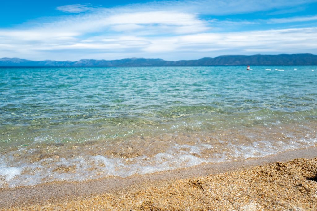Waves gently lap the sandy shore, with distant mountains under a bright blue, partly cloudy sky.