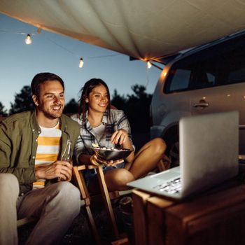 Two people sit under a canopy, watching a laptop, with string lights and a car visible in the evening outdoor setting.