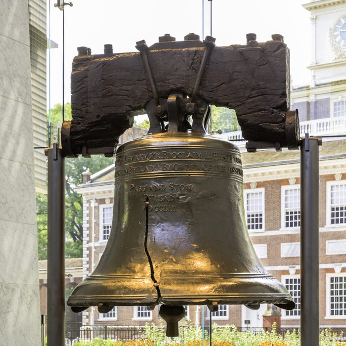 A large, cracked bell hangs, inscribed with historical text, in a museum setting with a historic building visible outside the window.