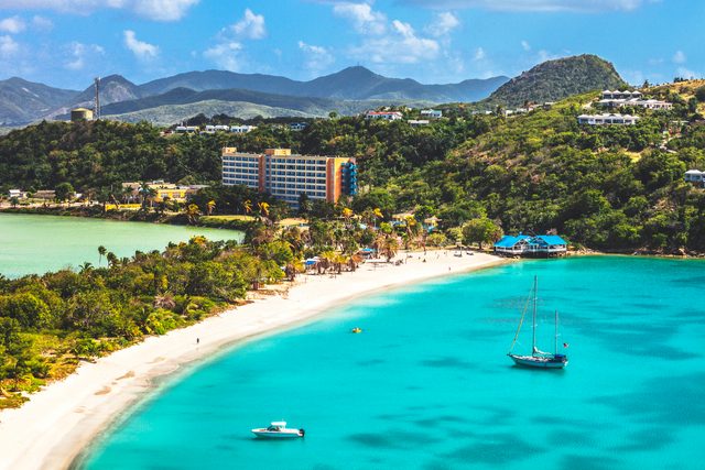 Sailboat drifts on turquoise waters near a sandy beach lined with trees, buildings, and hills in the background under a vibrant blue sky.