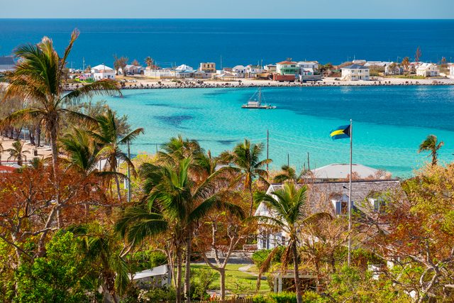 Palm trees sway over turquoise waters with a sailboat, quaint houses line the distant shore, and a flag flutters on a flagpole.