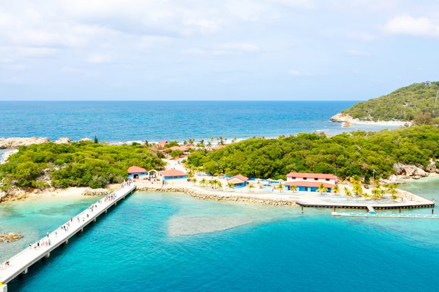 People walk along a pier leading to a small, lush island with colorful buildings, bordered by clear turquoise ocean under a partly cloudy sky.
